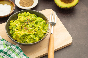 Fresh guacamole on a dish placed on a cutting wooden board with ingredients for homemade guacamole avocados, lemon, salt, and pepper. Top view.  Concept of traditional Mexican preparation