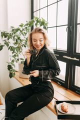 A cheerful happy sleepy young woman in black comfortable pajamas alone enjoys relaxing having breakfast drinking coffee in bed in a bright bedroom in a cozy house on a weekend morning