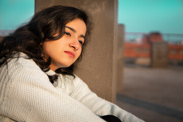 Outdoor portrait of an Asian, Indian beautiful, sad, serene young woman sitting alone near concrete pole on a roof top at day time and looking away while thinking with a blank expression at day time.