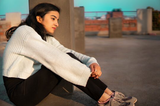 Outdoor full length image of an Asian, Indian beautiful, sad, serene young woman sitting alone near concrete pole on a roof top at day time and looking away while thinking with a blank expression. 