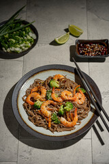 Buckwheat noodles with shrimps, carrots, broccoli, green onions and soy sauce. Vegan soba noodles. Asian food on grey textured table. Seafood healthy diet concept, copy space. 