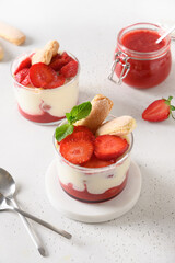 Layered strawberries dessert in glass jars with cookie savoyardi, mascarpone and whipped cream. Close up. Italian tiramisu with summer vibes on white.