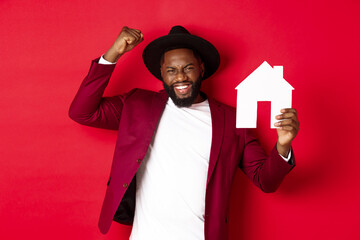 Real estate. Happy Black man celebrating, buying new house, showing paper home model and raising...