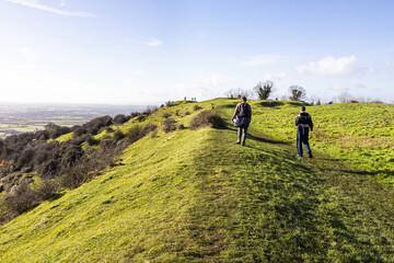Obraz premium Hikers walking along the rampart of a slight univallate Iron Age hillfort on Ring Hill at the Cotswold viewpoint of Haresfield Beacon, Gloucestershire UK