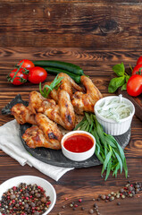 Grilled chicken wings on a wooden board with seasonings, basil, tomatoes and tarragon on a dark wooden background with sauce