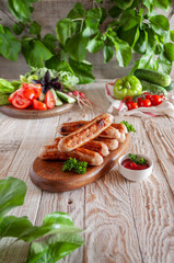 Grilled sausages on a cutting board on a light background with tree leaves, tomatoes, peppers, cucumbers and basil