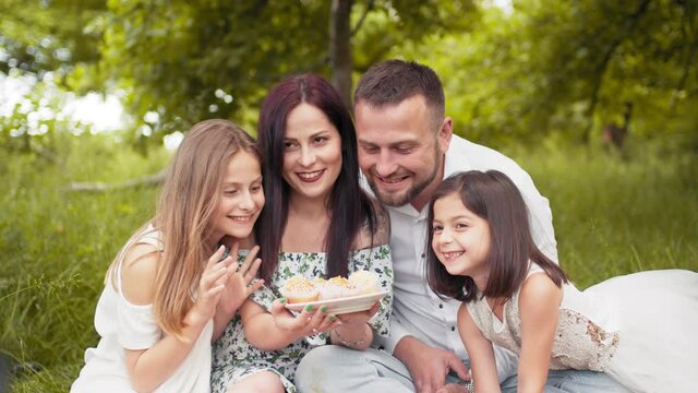 Young parents with two kids tasting delicious cupcakes while sitting together on soft blanket outdoors. Concept of family, relaxation and enjoyment. Happy family eating tasty cupcakes on picnic