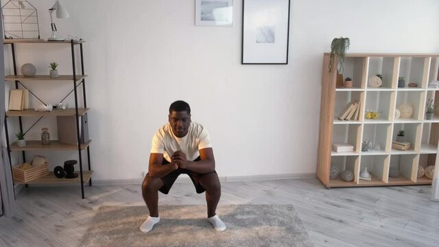 Home sport. Casual black man. Lockdown fitness. Healthy body. Handsome african guy in dark shorts white t-shirt doing squats workout in light room interior.