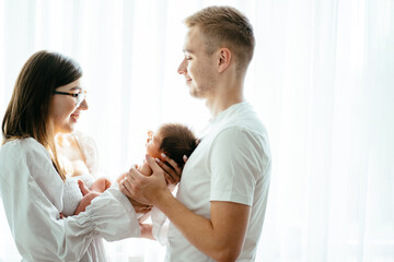 Fototapeta premium Young happy parents wearing white clothes holding their little baby girl, looking at her and smiling. Backlight emotional family lovely moment. Parent love, tenderness concept.