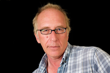 Obraz premium Studio portrait of blond haired blue eyed man in eyeglasses, front view, looking at viewer in front of black background, wearing worn plaid shirt.