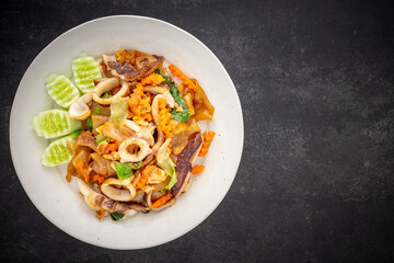 Pad See Ew Pla Muek, Thai food, stir fried soy sauce noodle with squid, egg, lettuce, carrot served with cucumber on dark tone texture background, top view