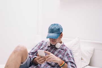 mature man with cap and lgtb bracelet lying on bed interacting with his cell phone
