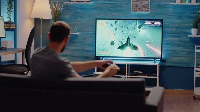 Young man with online console playing video games indoors while holding controller for action competition. Person using television and joystick for virtual electronic entertainment