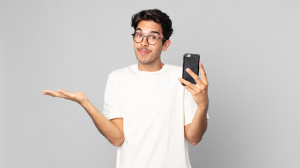 young hispanic man feeling puzzled and confused and doubting and holding a smartphone