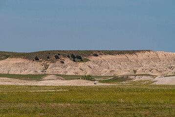 Badlands National Park, SD, USA - June 1, 2008: Landscape with beige geologic deposits appearing from under prairie on horizon under blue sky. Green plains up front.