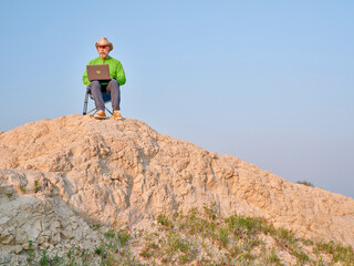 Senior man in cowboy hat is sitting on a folding chair and working on laptop in the middle of nowhere, early morning in the badlands of Pawnee National Grassland in Colorado