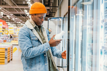 Positive African-American guy in denim jacket and orange hat takes milk bottle from fridge case in...