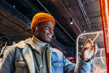 Smiling African-American man in warm denim jacket with wireless earphones uses self-service kiosk...