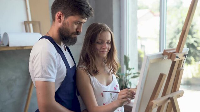 Couple of lovers painters collaborating in an art studio creating a masterpiece. Bearded artist teaches his girlfriend to paint oil painting canvas, use abstract artwork work in art workshop.