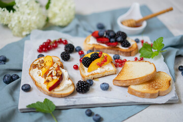 Toasted bread with cream cheese, honey, nuts, fruits and fresh berries. Summer tasty breakfast - dessert on white textured table. Selective focus with copy space.