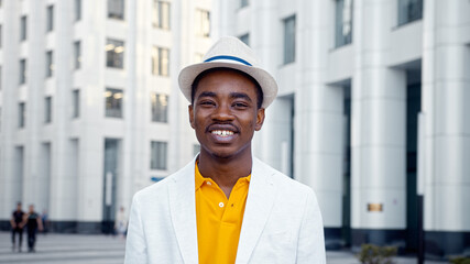 Smiling man businessman in white fedora hat with blue strip walks along city square with highrise...