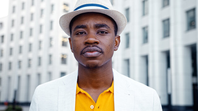 Serious Black Man In Yellow T Shirt Under White Jacket Looks Into Camera Standing By City Buildings