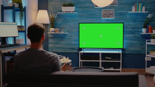 Caucasian person looking at green screen tv in living room. Man watching chroma key blank display using mockup template background in cozy living room eating pop corn late at night