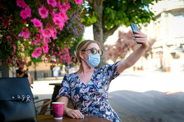 Beautiful young woman in a cafe in a sunglasses and medical mask and makes a selfie on the phone. A girl in a cafe on the street communicates online on a smartphone. Pandemic covid-19, quarantine.