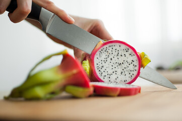 Female hands is cutting a dragon fruit or pitaya with pink skin and white pulp with black seeds on wooden cut board on the table. Exotic fruits, healthy eating concept