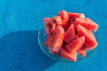 Chunky watermelon in a glass bowl on a blue painted wooden background, with direct sunlight.