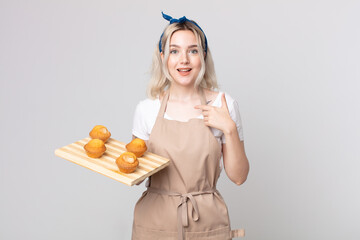 young pretty albino woman feeling happy and pointing to self with an excited with a muffins tray