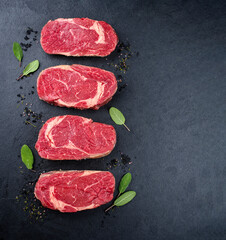 Four raw dry aged wagyu rib-eye beef steaks with herb and black salt offered as top view on a black board with copy space left