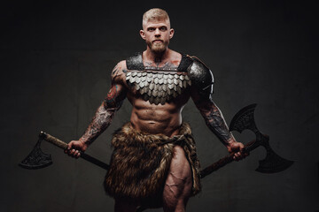 Brutal tattooed warrior wearing light armour and fur holding axes in dark studio