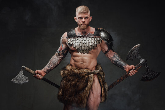 Strong tattooed warrior in light armour and fur with two axes in dark