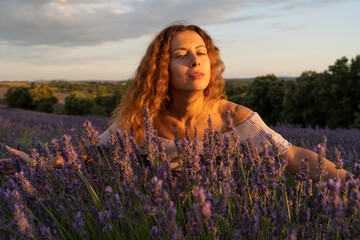 A young woman with curly hair in an off-the-shoulder dress enjoys the scent of lavender at sunset in a lavender field. Valensole, France 2021.