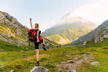 Caucasian and young person with his back turned, with a backpack and on top of a rock lifting one leg and one arm. Happy white woman hiking up the mountain