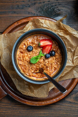 Homemade creme brulee with strawberries and mint in a metal bowl with a spoon on parchment on a dark brown wooden table. Top view.