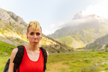Naklejka premium Young Caucasian girl with punk aesthetics cracking up and pretending to be sad with a mountain landscape in the background. tired hiker.