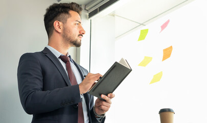 Portrait of concentrated male with eyeglasses writing on notebook while looking at sticky notes on glass wall in office. Businessman focused on creative business activity, developing business plan