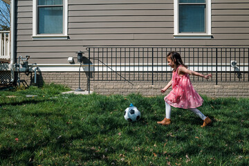 Little girl in pink dress kicking soccer ball in back yard