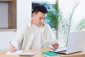 Fototapeta premium Young Asian man doing paperwork at home sitting at a desk, working on a laptop, focused on paying bills