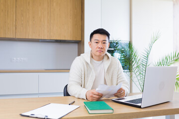 young Asian man counts bills, bank checks, loans or utilities while sitting at home at a kitchen room table. guy with laptop and calculator understands documents. pensive male became confused payments