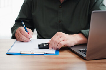Fototapeta premium Car insurance staff writing on the white paper clip file board hold car key and memo insurance data form computer laptop glasses on table : car insurance office work concept