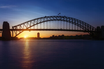 Sun setting behind the iconic Sydney Harbour Bridge in Sydney Harbour late in the evening in Australia with the silhouette of the city of North Sydney in the background.