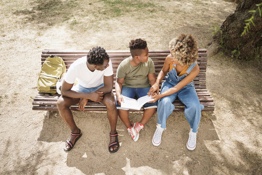Top down view of African American family doing school homework sitting on a bench in the park. Little son doing school work with help of his parents