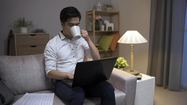 asian businessperson working overtime is looking at the computer screen with a thoughtful look on face while drinking tea on the sofa in the living room at home