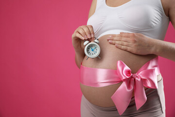 Young pregnant woman with alarm clock and bow on pink background, closeup. Time to give birth