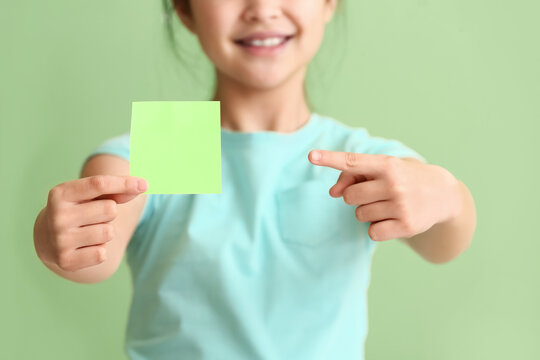 Little girl pointing at blank paper note on color background, closeup