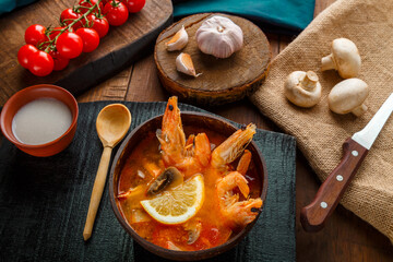 Tom yam soup with shrimps and coconut milk on a table on a black board near ingredients and a spoon.