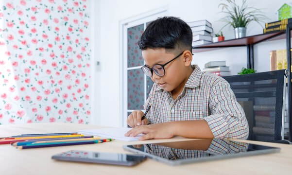 Smart Asian boy wearing glasses and grey checked shirt sitting on studying desk of tablet, mobile phone, and colorful pencils in reading room and concentrate on drawing on white paper